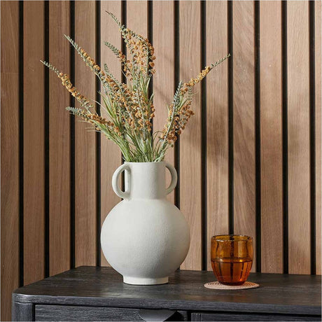 Matt white aluminium vase with two handles, 24cm, holding dried grasses on a dark table against a wood slat wall, with an amber glass nearby.