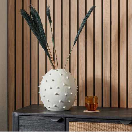 Matt White Aluminium Vase with Bobbles, 26cm round textured vase with dried plumes, styled on a dark sideboard beside an amber glass tumbler.