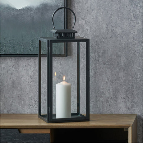 Black stainless steel and glass rectangular lantern with arched carry handle, holding a lit white pillar candle, shown on a wooden sideboard against a grey wall.