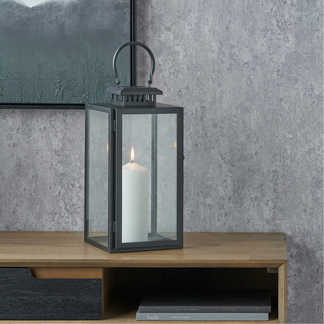 Black stainless steel and glass rectangular lantern, 39cm, with a white pillar candle inside, shown on a wooden sideboard against a grey textured wall.