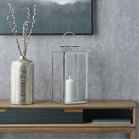 Silver stainless steel and glass rectangular lantern with an arched handle and lit white candle inside, displayed on a wooden console table beside a hammered metal vase.