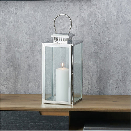 Silver stainless steel and glass rectangular lantern with an arched handle, holding a lit white pillar candle on a wooden table against a grey wall.