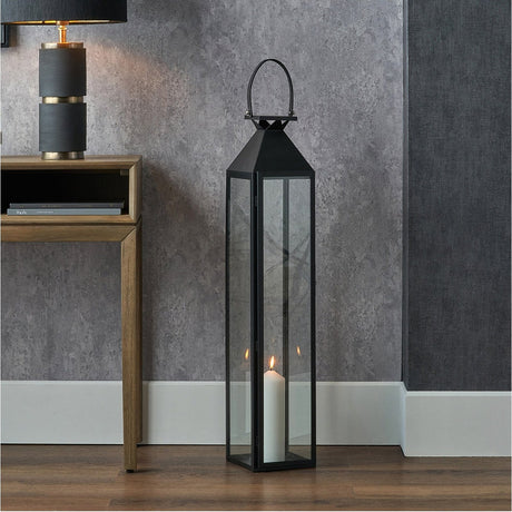 Black stainless steel and glass tall lantern, 93cm, with pyramid top and carry handle, holding a lit pillar candle, shown beside a wooden console table.