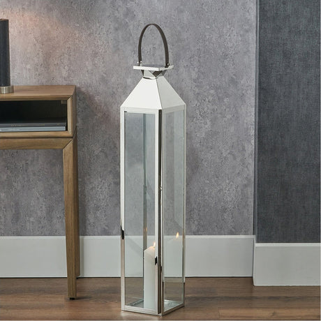 Tall silver stainless steel and glass lantern candle holder, 93cm, with a pointed top and rope handle, shown lit on a wooden floor beside a console table.
