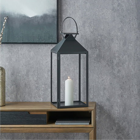 Black stainless steel and glass large lantern, 68cm, with pyramid roof and top handle, shown lit with a pillar candle on a wooden console table against a grey wall.