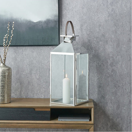 Silver stainless steel and glass large lantern with a leather handle and lit pillar candle inside, shown on a wooden console table against a grey textured wall.