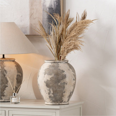 Lulia Antique Finish Stoneware Urn Vase, 31cm, rounded body in mottled grey and cream, holding dried pampas grass on a sideboard beside a stone-effect table lamp.