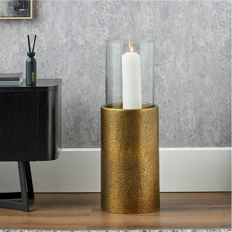 Antique brass hurricane lamp with clear glass shade and lit white pillar candle, styled on a wooden floor beside a dark cabinet.
