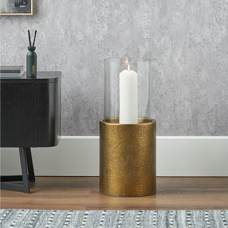 Antique brass hurricane lamp, 54cm, with clear glass cylinder holding a lit white pillar candle, shown on a textured cylindrical base beside a dark cabinet in a living room setting.