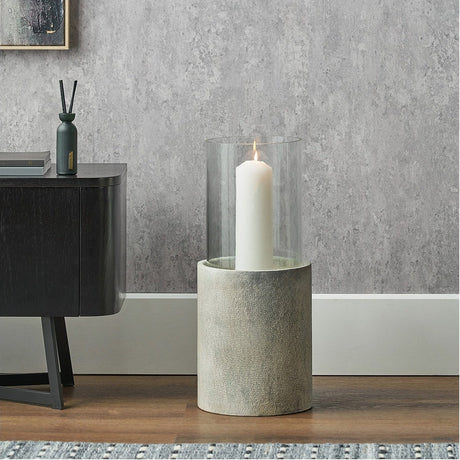 Stone Grey Aluminium & Clear Glass Hurricane Lamp, 53cm, with a lit white candle, styled on a wooden floor beside a dark cabinet against a grey textured wall.