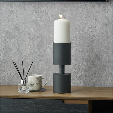 Matt black aluminium pillar candle holder, 30cm, holding a lit white pillar candle on a wooden sideboard with a diffuser and marble mug nearby.