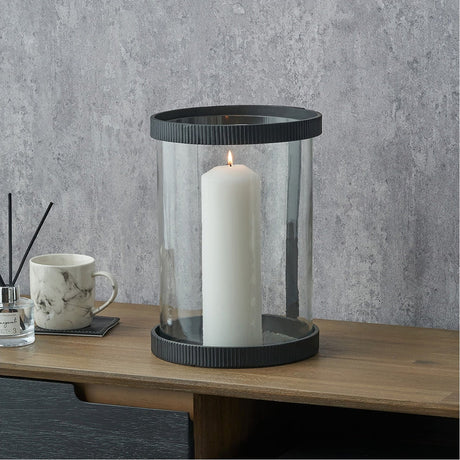 Black Ribbed Metal & Clear Glass Large Hurricane Lamp, 31cm, holding a lit white pillar candle, styled on a wooden sideboard beside a marble mug against a grey wall.