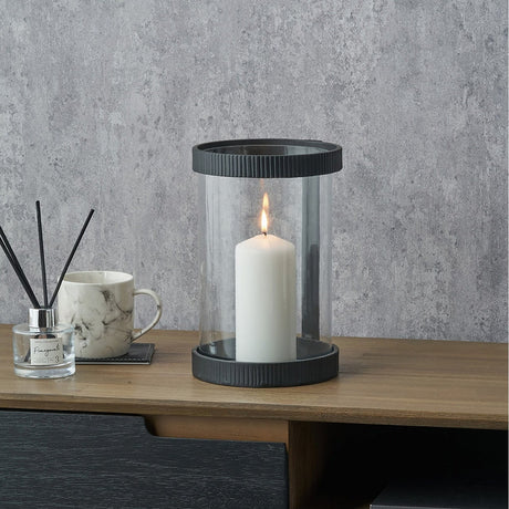 Black Ribbed Metal & Clear Glass Hurricane Lamp, 25cm, with a lit white pillar candle inside, shown on a wooden sideboard against a grey textured wall.