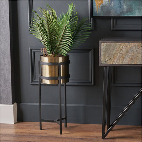 Brass and matt black floor planter, 66cm, with cylindrical brass pot on a black metal stand, styled with faux palm fronds beside a dark panelled wall.