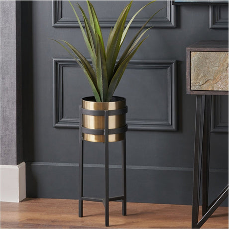 Brass and matt black floor planter, 52cm, on a tall black metal stand, holding a faux plant against a dark panelled wall.