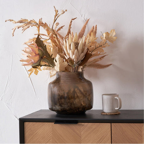 Cerdanya Brown & Beige Glass Vase, 23cm, holding dried floral stems on a black sideboard with a mug, set against a white wall.