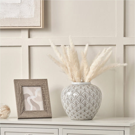 Barbier White Art Deco Fan Ceramic Vase, 22cm, with scalloped patterned body, holding dried pampas grass on a sideboard beside a framed picture.