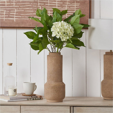 Gaiana Concrete Brown Stoneware Vase, 37cm, holding green foliage and white hydrangea stems on a wooden sideboard beside a candle, mug, and books.