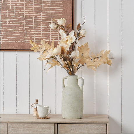 White textured ceramic vase with two handles, 35cm, holding faux magnolia stems and autumn leaves on a wooden sideboard against a white panelled wall.