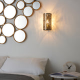 Endon Cliffmere antique brass wall light with cut-out secret-garden pattern, illuminated beside a bed with a circular gold-framed mirror display.