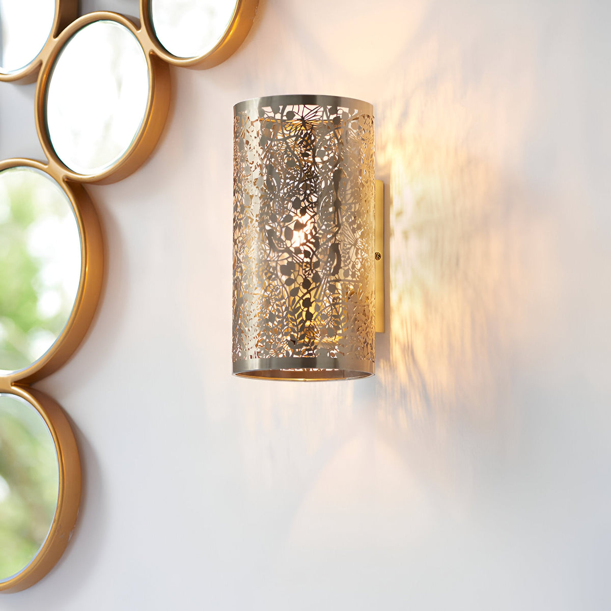 Endon Cliffmere wall light in antique brass, cylindrical shade with cut-out leaf, dragonfly and bird design, mounted beside a gold circular mirror.