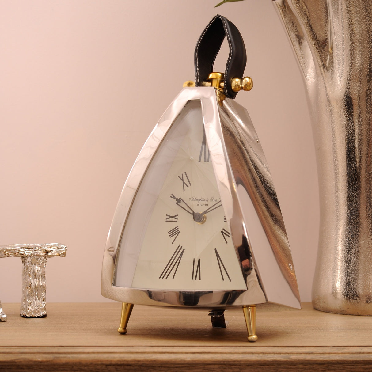 Libra Interiors Polished Nickel Curved Front Mantel Clock with cream roman numeral dial, black leather loop handle and brass feet on a wooden sideboard.