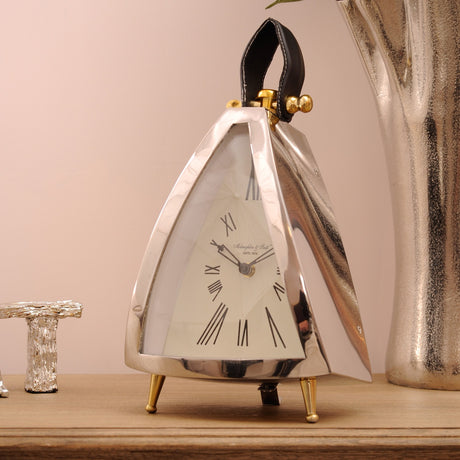 Libra Interiors Polished Nickel Curved Front Mantel Clock with cream roman numeral dial, black leather loop handle and brass feet on a wooden sideboard.