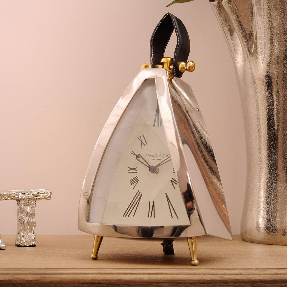Libra Interiors polished nickel triangular mantel clock with cream roman numeral dial, black leather loop handle and splayed brass feet on a wooden sideboard.