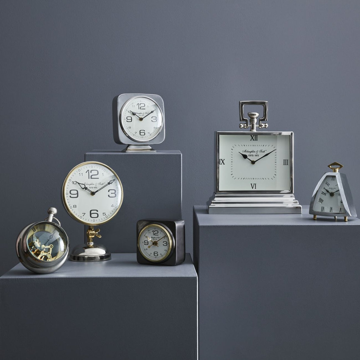 Libra Interiors Polished Nickel Curved Front Mantel Clock display with assorted silver and brass table clocks arranged on grey plinths against a dark grey wall.