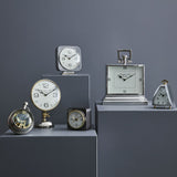 Libra Interiors Polished Nickel Curved Front Mantel Clock display with assorted silver and brass table clocks arranged on grey plinths against a dark grey wall.