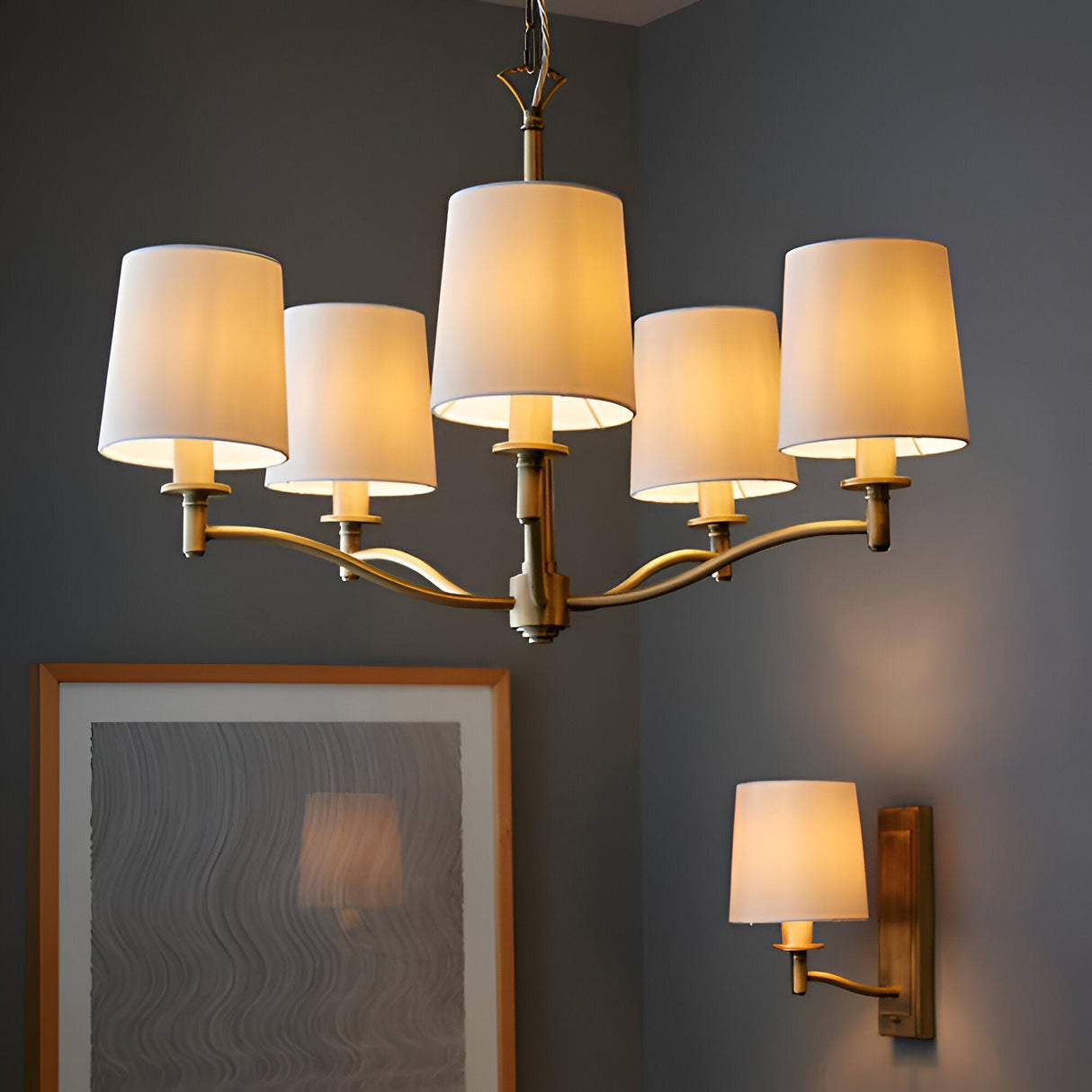 Endon Castwell 5 light pendant in antique brass with vintage white fabric shades, shown lit above a framed picture with a matching wall light on grey walls.