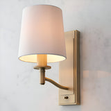 Endon Castwell wall light with matt antique brass bracket, toggle switch and vintage white faux-silk tapered shade, mounted on a light background.