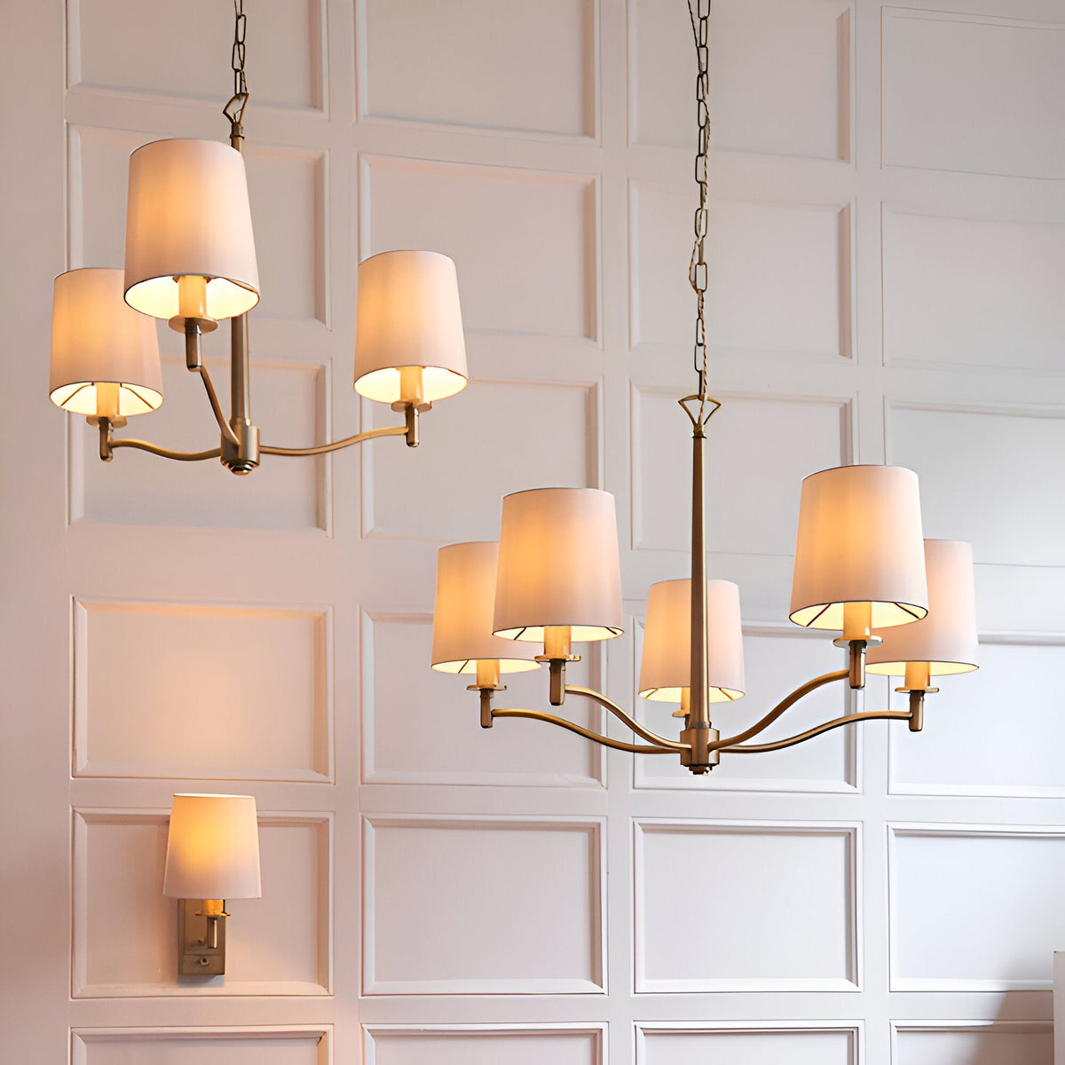 Endon Castwell wall light in antique brass with a vintage white faux-silk shade, shown alongside matching chandeliers on a panelled wall.
