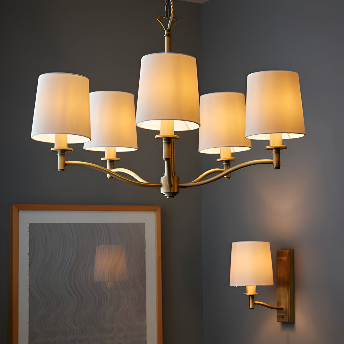 Endon Castwell chandelier and matching wall light in antique brass with vintage white faux-silk shades, shown above a framed artwork on a grey wall.