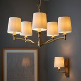 Endon Castwell chandelier and matching wall light in antique brass with vintage white faux-silk shades, shown above a framed artwork on a grey wall.