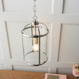 Endon Caldbury pendant light in satin nickel with a cylindrical clear glass lantern shade and single E27 filament lamp, hung from a chain against panelled wall.