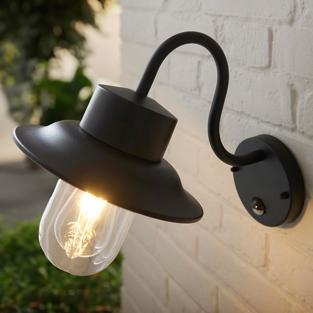 Endon Bevfield fisherman-style outdoor wall light in textured black with clear glass diffuser, lit bulb and built-in PIR sensor, mounted on a white brick wall.