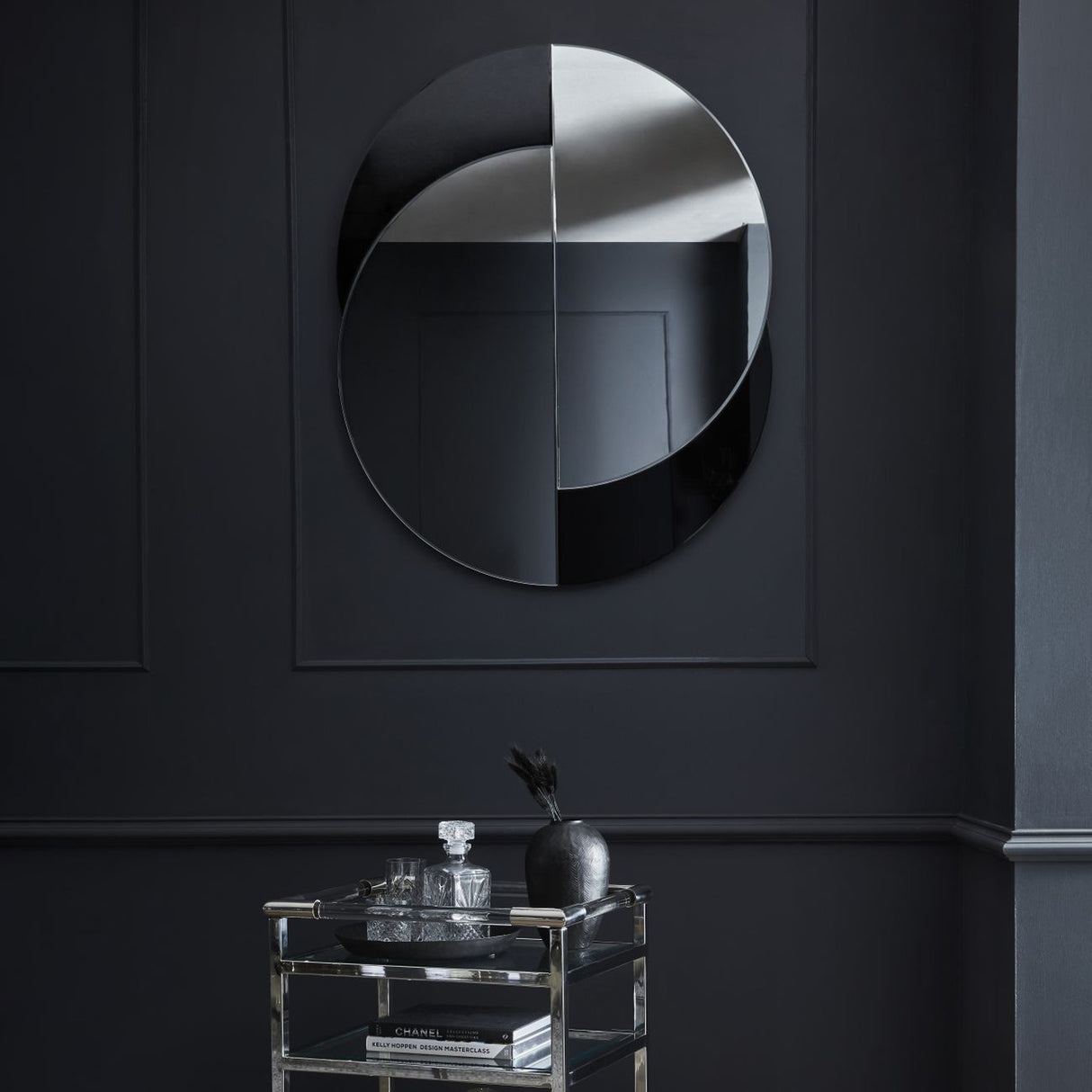 Libra Interiors Overlapping Circles wall mirror in clear and black glass, mounted on a dark panelled wall above a chrome and glass drinks trolley.