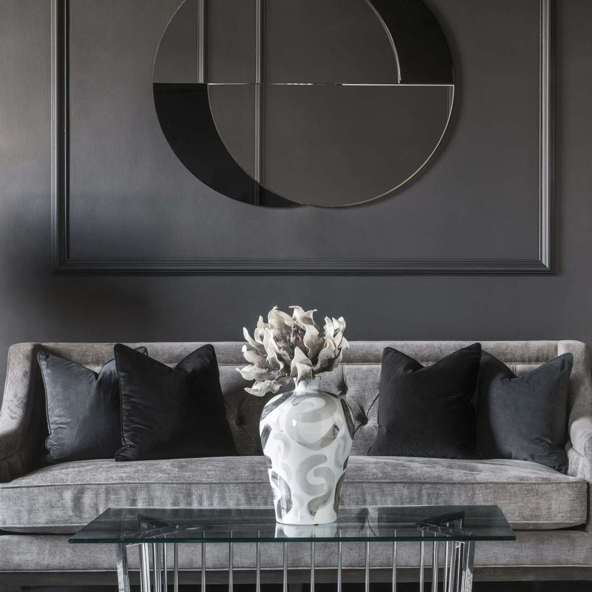 Libra Interiors Overlapping Circles wall mirror in clear and black glass above a grey sofa with dark cushions and a decorative vase on a glass table.