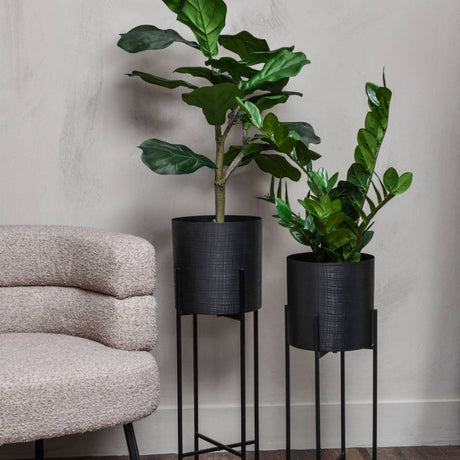 Libra Interiors Etched Iron Set of 2 Floor Standing Planters in matt black, holding leafy greenery on slim four-leg stands beside a boucle armchair.