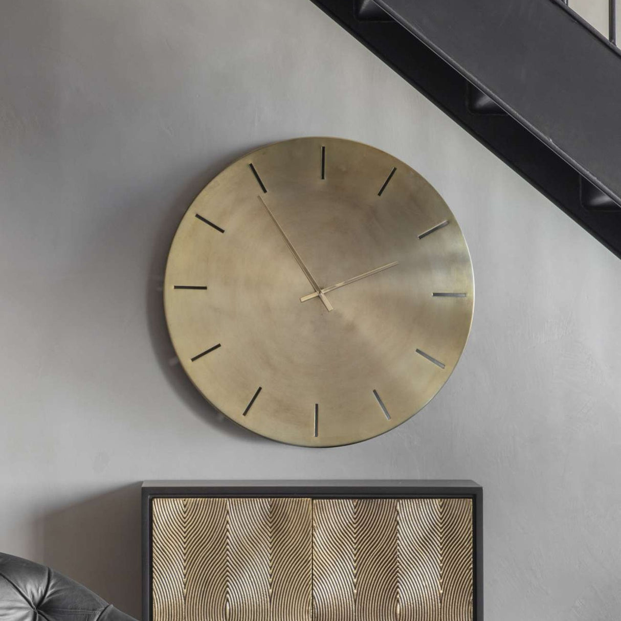 Libra Interiors Brushed Gold Iron Round Wall Clock with raised hour markers and slim hands, mounted on a grey wall above a patterned gold sideboard.