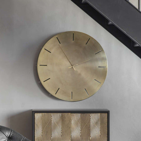 Libra Interiors Brushed Gold Iron Round Wall Clock with raised hour markers and slim hands, mounted on a grey wall above a patterned gold sideboard.