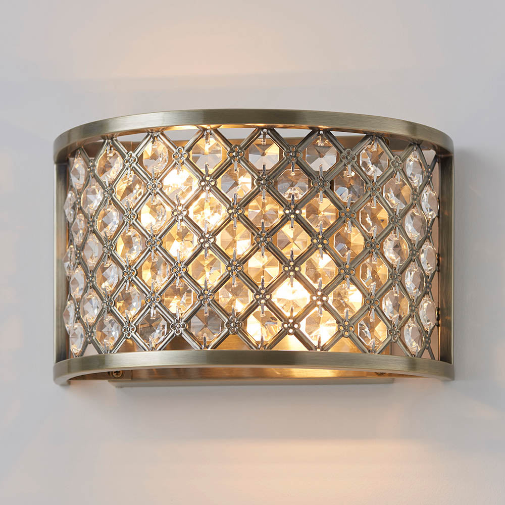 Endon Prescott 2-light wall light in antique brass with a curved steel lattice frame set with clear crystal beads, illuminated against a white wall.