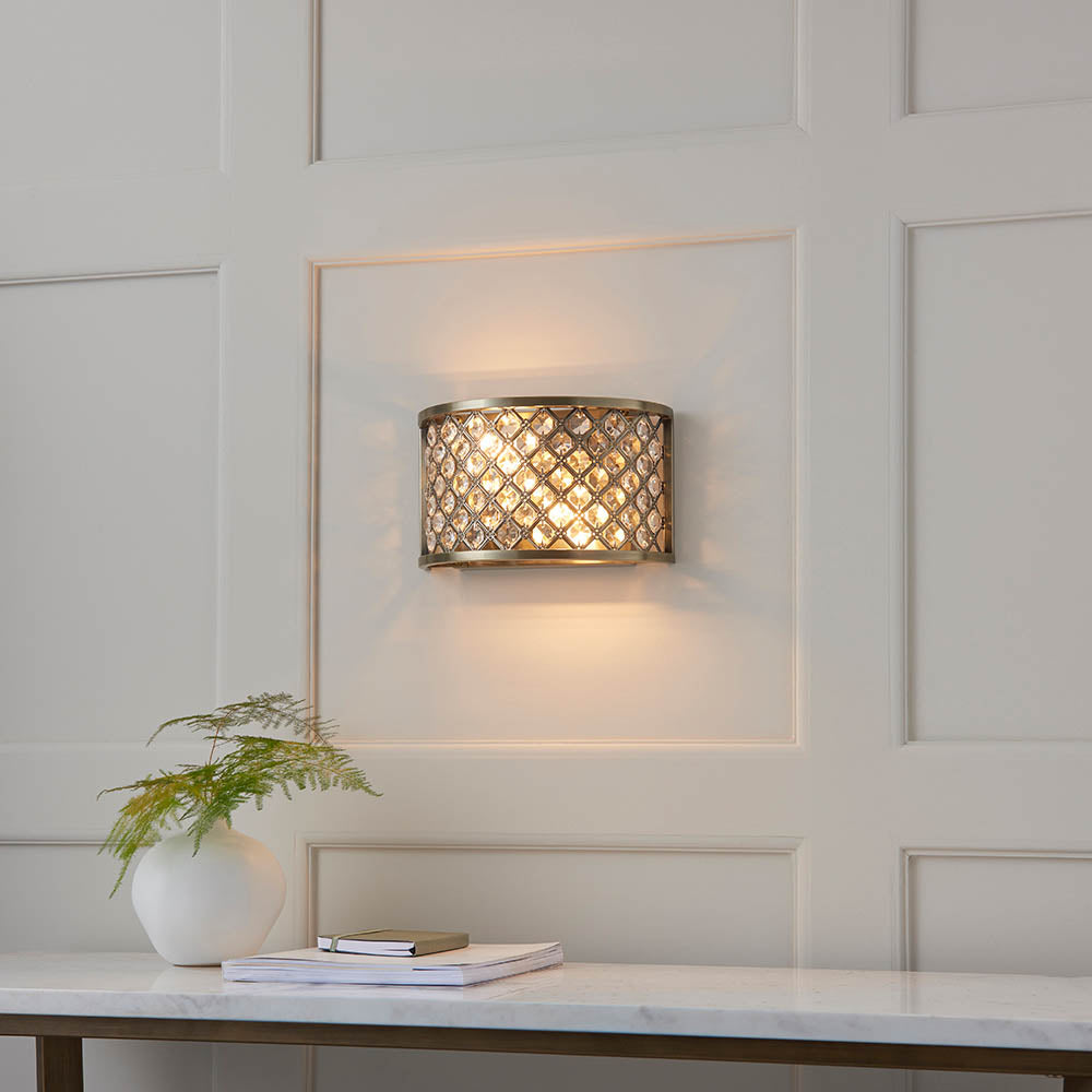 Endon Prescott two-light wall fitting in antique brass with crystal beads, mounted on a panelled wall above a marble console with a fern and books.