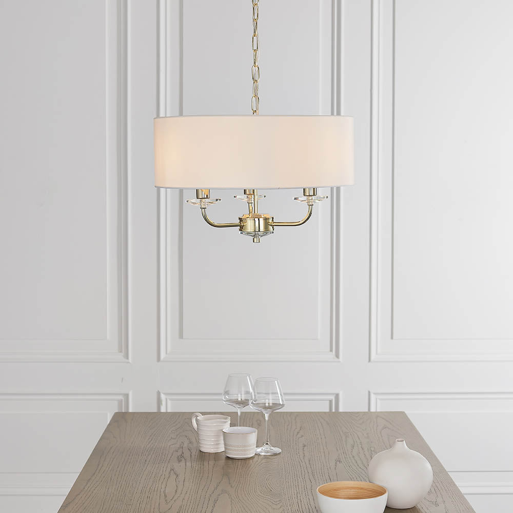 Endon Castdon 3 light pendant in brass with white faux silk drum shade and crystal detailing, suspended by chain above a wooden dining table.