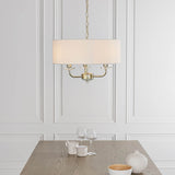 Endon Castdon 3 light pendant in brass with white faux silk drum shade and crystal detailing, suspended by chain above a wooden dining table.
