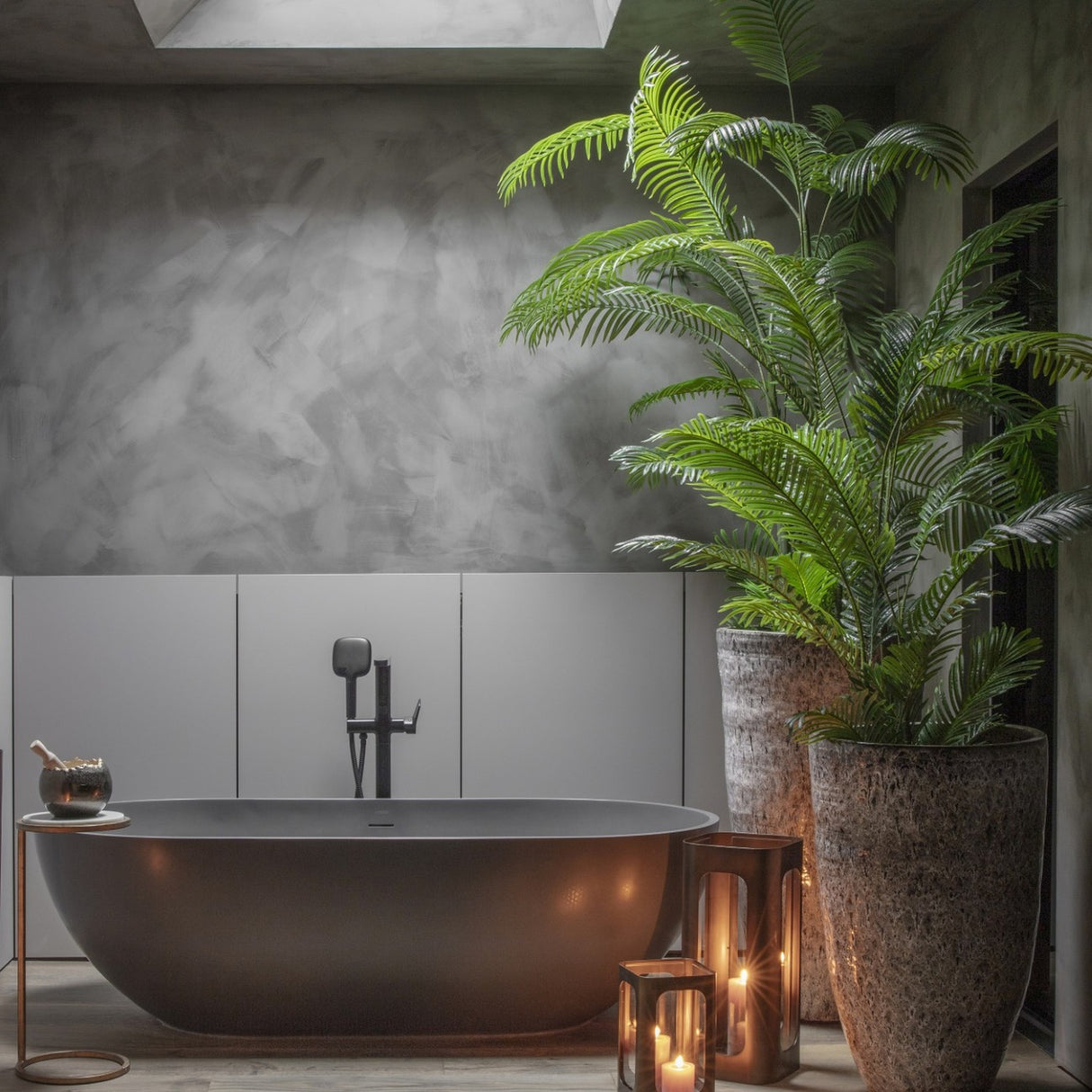Libra Interiors Faux Areca Palm in tall textured stone-effect planters beside a matte black freestanding bath, candle lanterns and grey plaster walls.