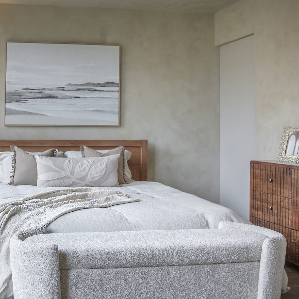 Libra Interiors Textured Boucle Storage Ottoman in cream, placed at the foot of a wooden bed with white bedding, beside a wood dresser in a neutral bedroom.