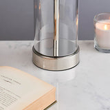 Endon Caldley table lamp detail showing the clear cylindrical glass base and polished nickel fitting on a marble surface beside an open book and candle.