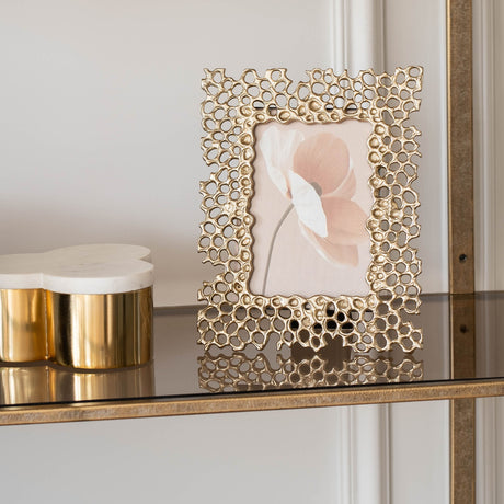 Libra Interiors Openwork Cast Aluminium Photo Frame in gold with honeycomb border, holding a floral print, styled on a glass shelf beside a marble box.